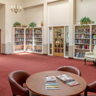 A cozy library reading room with bookshelves, armchairs, and a round table with magazines.