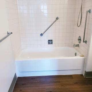A bathroom featuring a bathtub with handrails and a tiled wall, complemented by a wooden floor.
