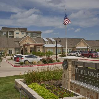 A welcoming entrance to a building complex featuring well-maintained landscaping, parked cars, and an American flag flying in the foreground.