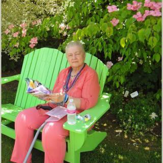 An elderly woman sitting on a green bench in a garden, reading.