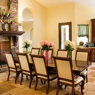 A cozy dining room with a large table surrounded by chairs, adorned with floral arrangements and featuring a stone fireplace.