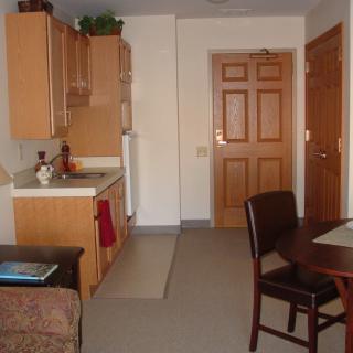 A compact apartment kitchen with a small table and chairs, showcasing wooden cabinets and a front door.