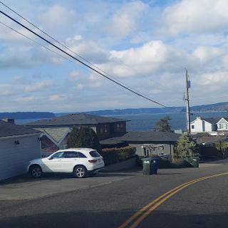 A scenic view of a winding road leading towards the seaside, showcasing houses and a white car parked nearby under a partly cloudy sky.