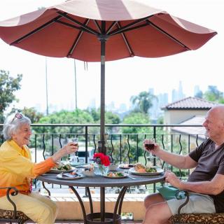 An elderly couple enjoying a meal and wine on a patio under an umbrella.
