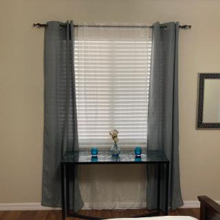 A room with a window featuring blue curtains, blinds, and a slim table adorned with decorative items.