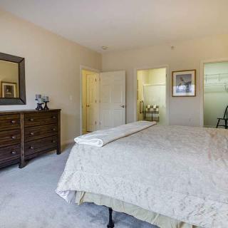 A cozy bedroom with a neatly made bed, a wooden dresser with a mirror, and soft carpet flooring.