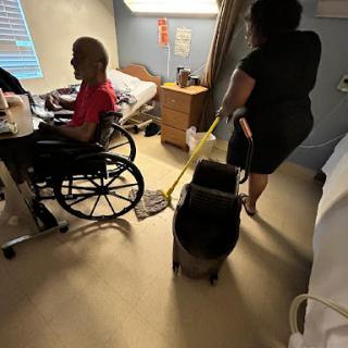 A caregiver cleans the floor in a hospital room while attending to a patient in a wheelchair.