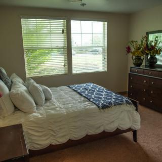 A cozy bedroom featuring a neatly made bed, a dresser with decorative items, and large windows letting in natural light.