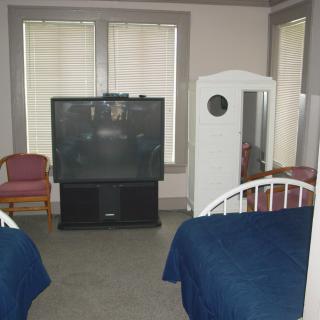 A bedroom with two beds, a large television, chairs, and a mirrored wardrobe.