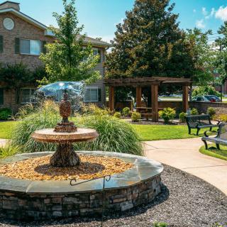 A tranquil garden setting featuring a stone fountain, benches, and lush landscaping with trees and a pathway.