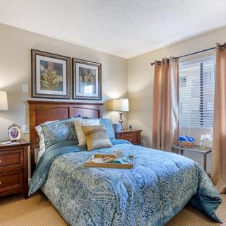 A cozy bedroom with a neatly made bed, flanked by nightstands and lamps, complemented by framed artwork above the headboard. Natural light filters through the window, enhancing the serene atmosphere.