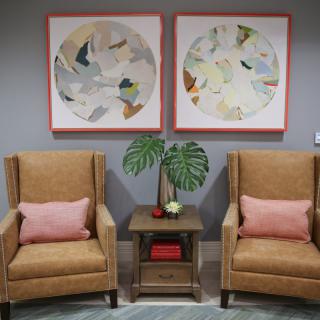 A cozy living room scene featuring two beige armchairs with pink pillows, a small side table with a plant, and abstract artwork on the wall.