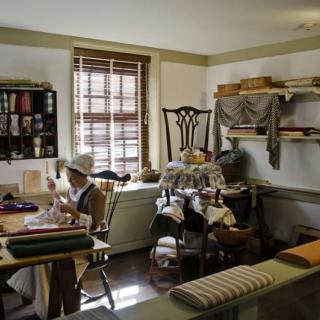 A historical sewing workshop with a person working on textiles amidst vintage decor.
