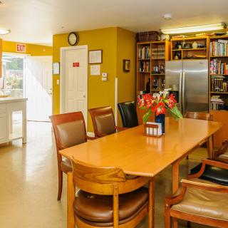 A cozy kitchen and dining area with a large wooden table, surrounded by chairs, adorned with flowers and flanked by bookshelves and a refrigerator.