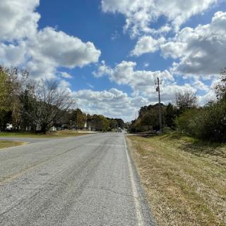 A rural road stretches into the distance under a partly cloudy sky, bordered by trees and grass.