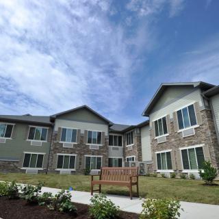 A modern residential building with a landscaped garden and wooden bench under a partly cloudy sky.