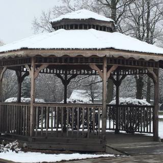 A wooden gazebo covered in snow, surrounded by winter trees.