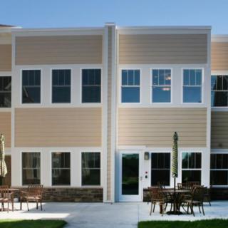 A modern two-story building with a beige exterior and large windows, featuring outdoor patio furniture.