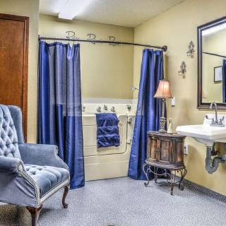 A cozy bathroom featuring a bathtub with blue curtains, a sink with a mirror above, and an upholstered chair.
