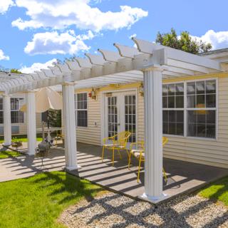 A sunny backyard featuring a white pergola attached to a house, with a patio area and green lawn.