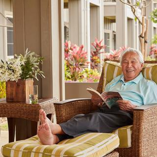 An elderly man is comfortably reading a book while relaxing on a patio chair.