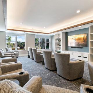 A modern living room with comfortable armchairs facing a large television, illuminated by natural light from the windows.