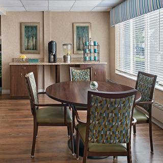 A cozy dining area with a round table surrounded by four patterned chairs, featuring large windows with blinds and decorative wall art.