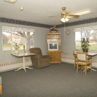 A cozy living room with a TV, two tables with chairs, and windows overlooking the outside.
