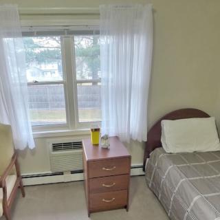 A simple bedroom with a single bed, chair, nightstand, and window with curtains.