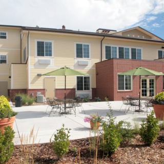 A modern building with a spacious outdoor patio featuring tables, chairs, and green umbrellas, surrounded by plants.