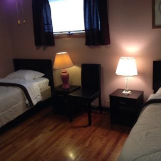 A cozy bedroom with two single beds, each accompanied by a nightstand and lamp, featuring dark curtains and a wooden floor.