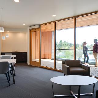 A modern office space with large glass walls, minimalist furniture, and people conversing on a balcony.