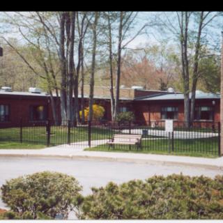 A view of a single-story brick building surrounded by trees and a fence, with bushes lining the driveway.
