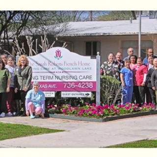 A group of healthcare workers posing in front of a nursing home with a sign and flowers.