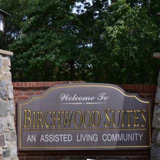 An outdoor sign for Birchwood Suites, an assisted living community, surrounded by stone pillars and greenery.