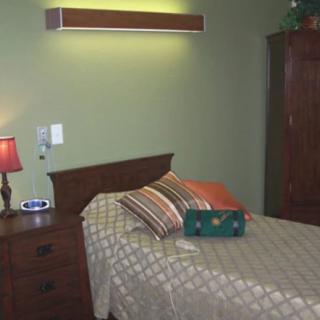A cozy bedroom featuring a single bed with striped pillows and a quilted bedspread, accompanied by a nightstand with a lamp and an armoire against the green-painted wall.
