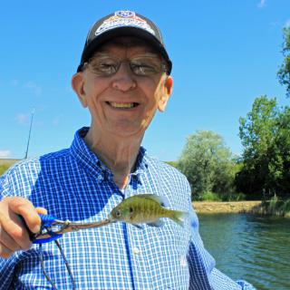 A man happily holding a small fish by a lake on a sunny day.