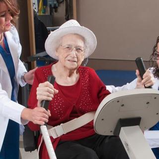 An elderly woman receiving assistance from healthcare professionals during a physical therapy exercise.