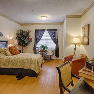 A cozy bedroom interior with a neatly made bed, small table and chairs by the window, and warm lighting.