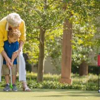 A senior person helps a child play golf outdoors on a sunny day.