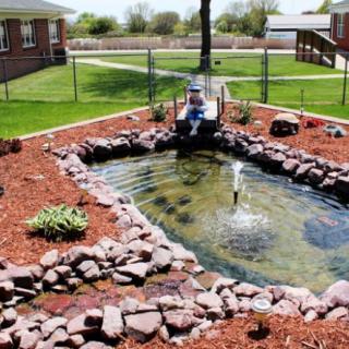 A beautifully landscaped garden pond with a small bridge and fountain, surrounded by rock borders and green grass.