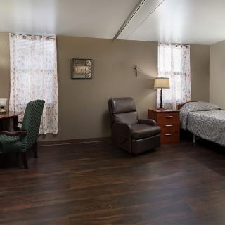 A neatly arranged bedroom with a bed, recliner, desk, and lamp.