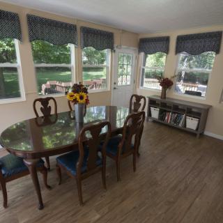 A bright dining room with a wooden table and chairs, adorned with a vase of sunflowers and surrounded by large windows.