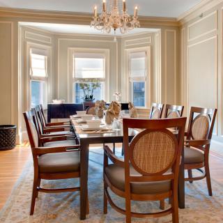 An elegant dining room with a long table and chandelier, surrounded by wooden chairs and large windows.