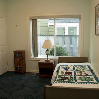 A simple bedroom with a single bed, quilted blanket, nightstand, and a lamp by the window.