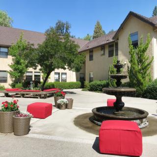 A sunny courtyard with red outdoor furniture and a central fountain, surrounded by buildings and greenery.