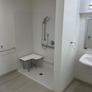 An accessible bathroom with a shower area featuring a folding chair and grab bars, alongside a sink and mirror.