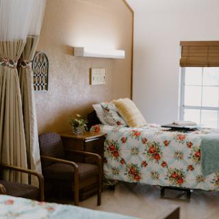 A cozy hospital room featuring two beds with floral bedding, a chair, and natural light streaming through the window.