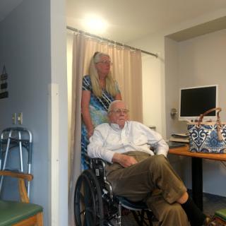 A woman stands behind an elderly man in a wheelchair, who is seated in a well-lit room with a computer nearby.