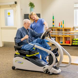 A healthcare professional assists a senior patient with exercise on specialized equipment during a rehabilitation session.